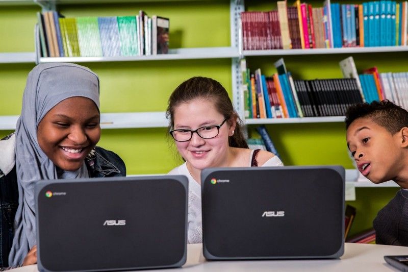 Three students sitting at a table and looking at one student's computer screen. 