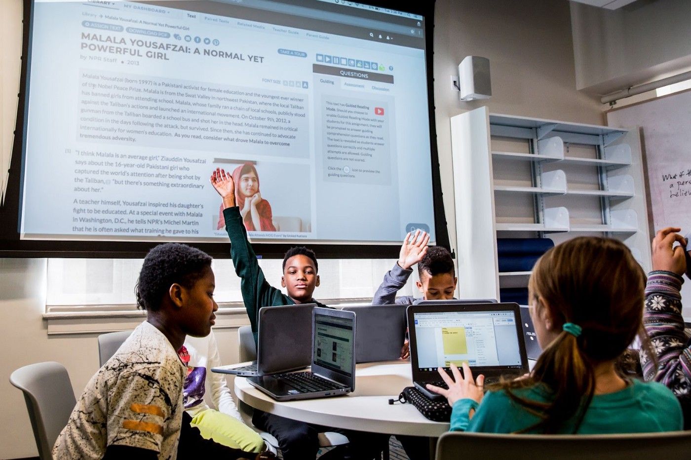 Students sitting around a round table with their laptops open. Some students are raising their hands. 