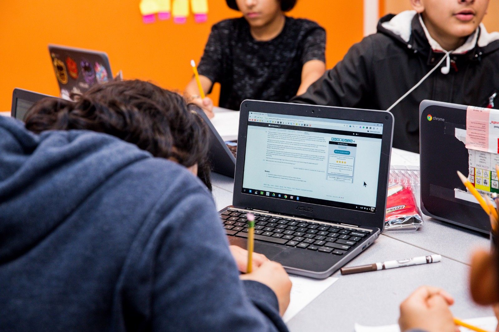 Several students sitting at a table, writing on paper with their computers open to CommonLit.org. 