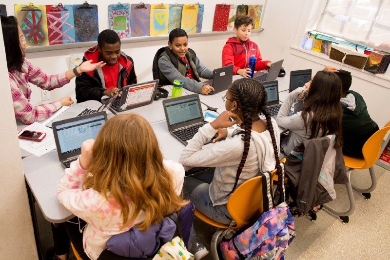 A group of students and their teacher sitting at a table, working on CommonLit.org on their computers. 