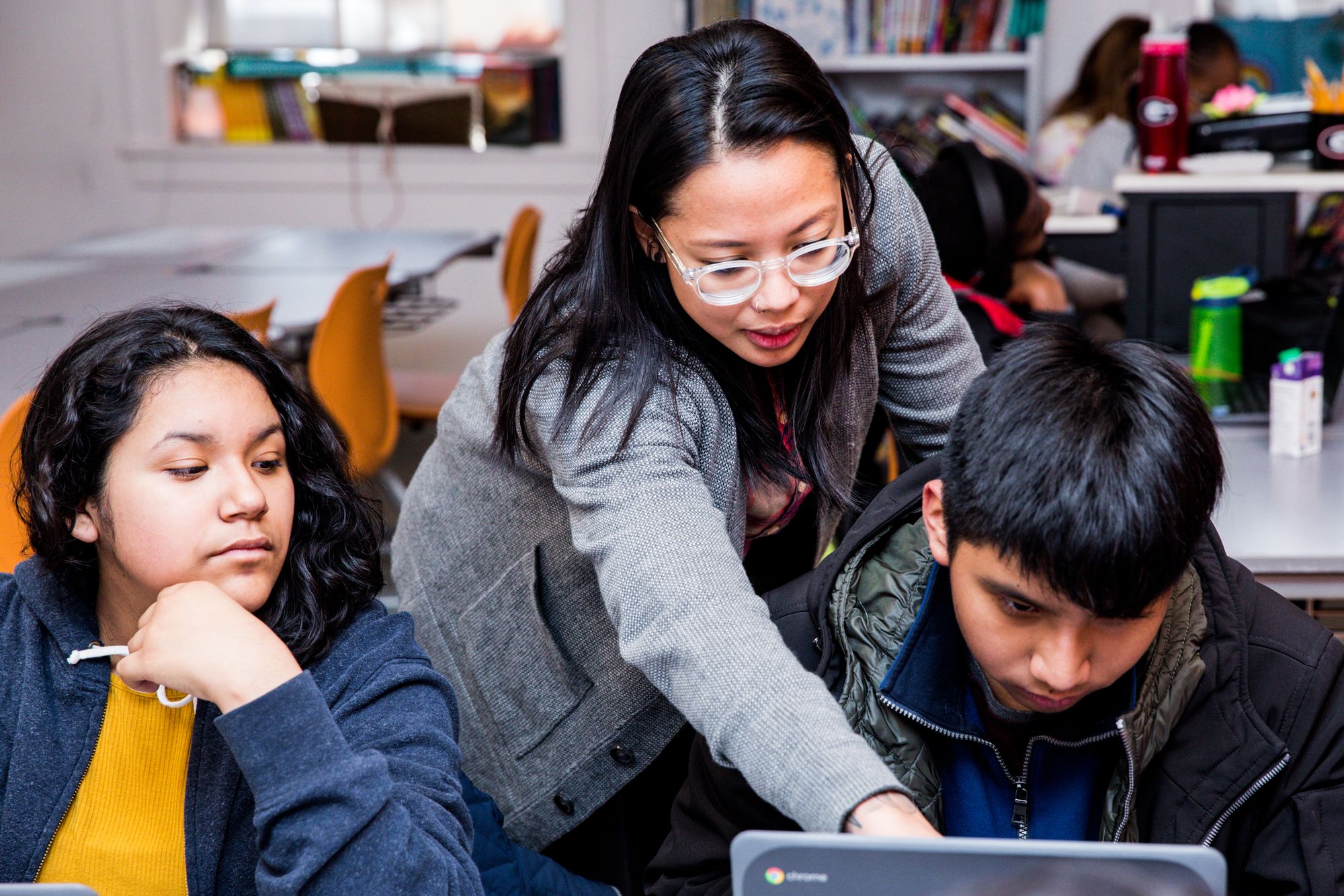 A teacher helps a student on his computer while another student looks on. 