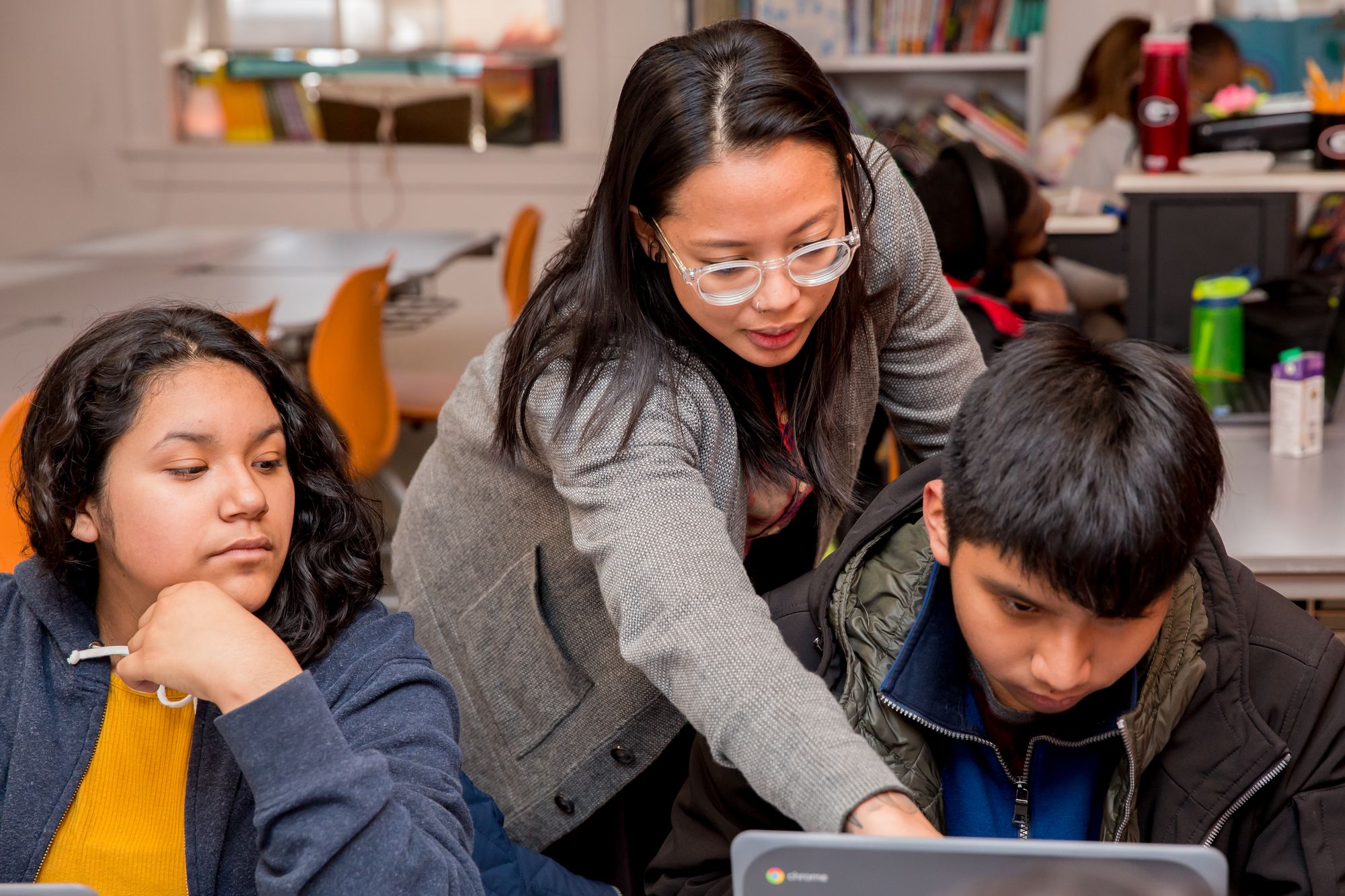 A teacher is standing behind two students, helping them with lessons on their computer.