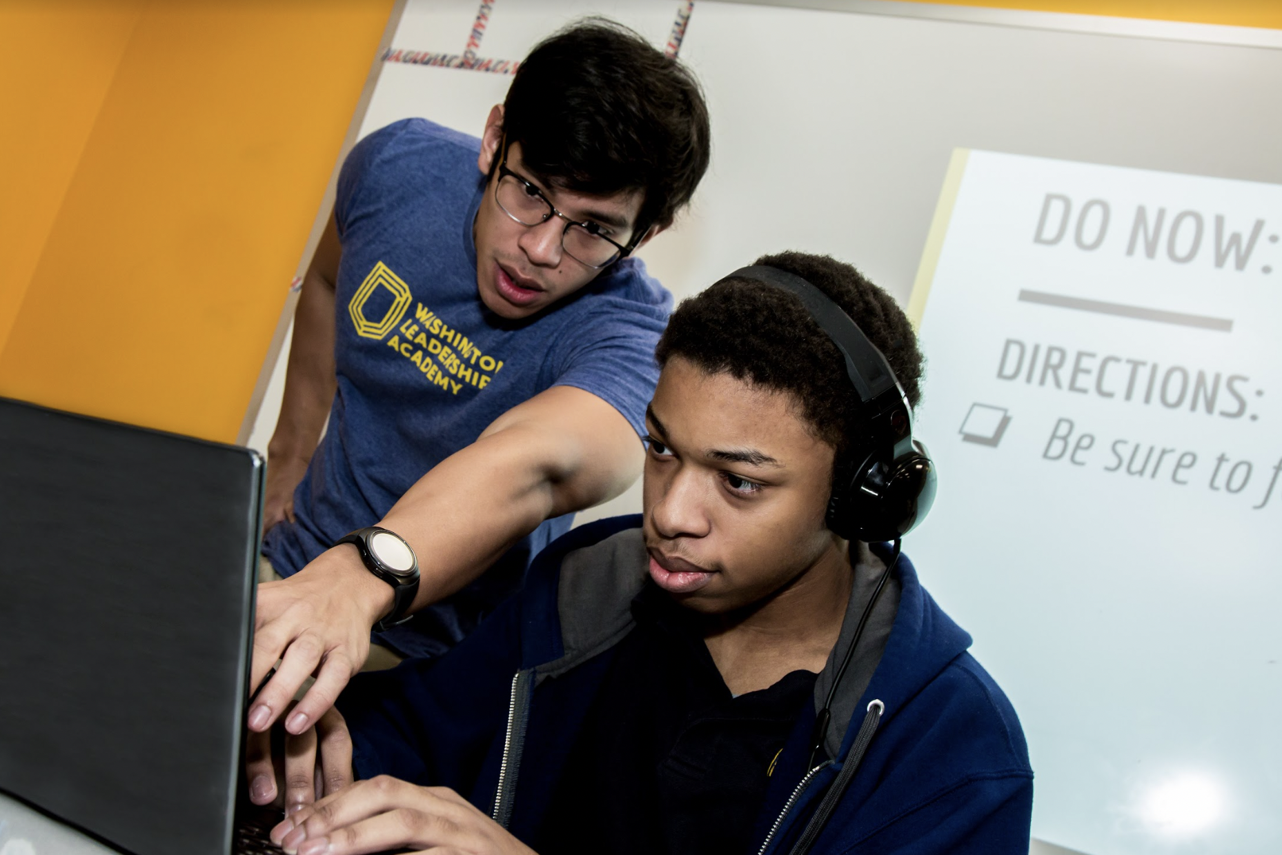 A teacher points at a student's laptop. The student is sitting and wearing headphones. 